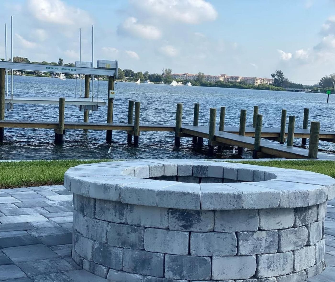 A stone fire pit on a waterfront lawn with a Mariner-built dock beyond it